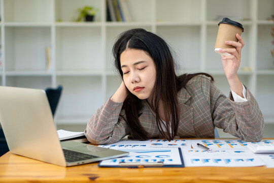 Sleepy Asian Businesswoman Yawning At Work In The Afternoon After Being Tired And Fed Up With Working With Laptop And Financial Graph Documents At Work.