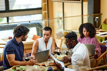 A group of casual businesspeople is sitting in a restaurant for a meeting, discussing a project while having dinner.