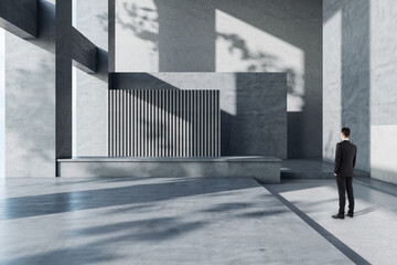 Businessman in suit back view looking at blank black banner with place for your logo or text on dark grey wall in sunlit spacious hall with concrete design, mock up