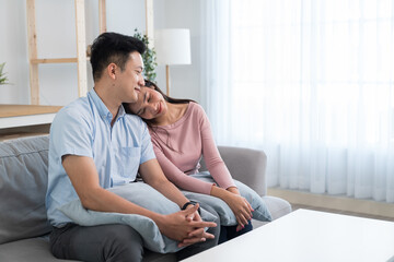 Asian young loving couple sit on sofa and hugging each other with love. Attractive romantic new marriage man and woman enjoy spend time together on anniversary valentines day in living room at home.