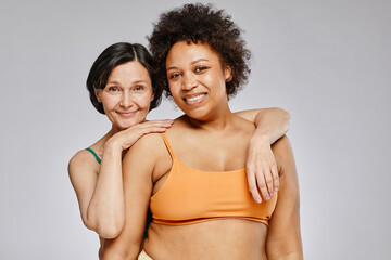 Minimal waist up portrait of two real women wearing underwear and smiling at camera against grey background