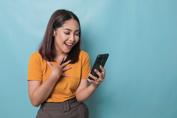 An excited young woman is smiling while holding her smartphone in her hand, isolated on blue background
