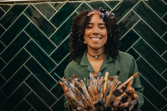 Portrait of young female artist holding dirty brushes and looking at camera and smiling in creative studio