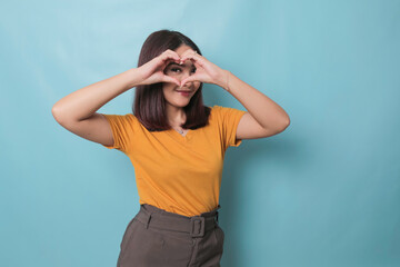 Attractive young Asian woman feels happy and romantic shapes heart gesture expresses tender feelings wears casual yellow t-shirt against blue background. People affection and care concept