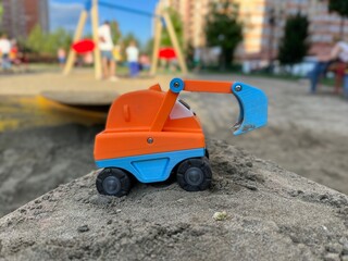 Vivid colorful orange toy excavator with big black wheels and a blue dipper (bucket) shot in the sandbox on a playground against blurry background