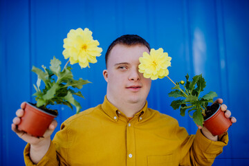 Happy young man with Down syndrome looking at camera and holding pot flowers against blue...