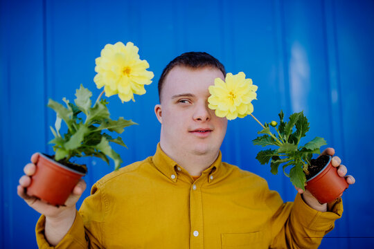 Happy Young Man With Down Syndrome Looking At Camera And Holding Pot Flowers Against Blue Background.