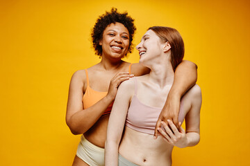 Naklejka premium Waist up portrait of two carefree young women dark skin and fair skin embracing while standing against vibrant yellow background in underwear
