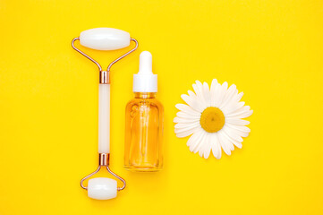 Serum bottle, jade facial massage roller and chamomile on a yellow background. Anti-aging, lifting...