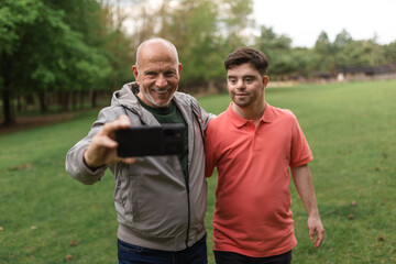 Happy senior father with his young son with Down syndrome taking selfie in park.