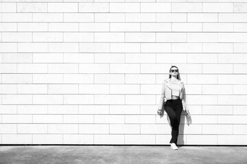 Lifestyle and fashion concept. Beautiful woman with blouse and jacket, black pants, white sneakers and sunglasses standing in front of huge brick wall background with copy space. Black and white image
