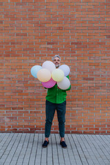 Fun portrait of happy energetic mature man holding balloons in street and hiding behind them, feeling free.