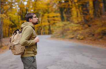 Obraz premium Young handsome man posing in autumn forest on road. young hipster guy with backpack , traveller standing in woods, Hiking, Forest, Journey, active healthy lifestyle, adventure, vacation concept.