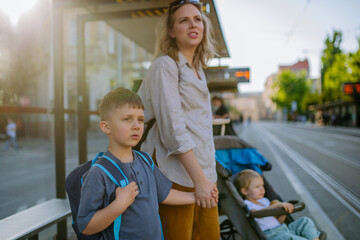 Young mother with little kids waiting on bus stop in city.