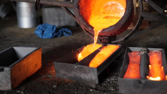Slow motion foundry bucket pouring orange metal into moulds with flames in a rustic factory setting.