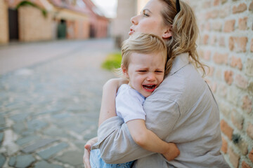 Mother consling her little daughter crying, holding her in arms in street in summer.