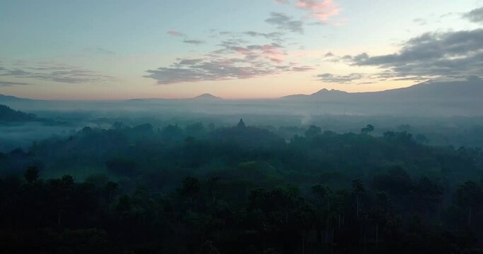 Aerial orbit flight of Borobudur Temple in Melang during hovering fog during golden morning - Panorama green landscape surrounding largest temple in world