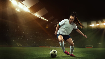 Female soccer, football player dribbling ball in motion at stadium during sport match over evening...