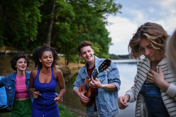 Fototapeta premium Group of young friends having fun on picnic near a lake, laughing, dancing and playing guitar.