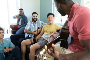 Multiracial happy male friends with beer bottles and cellphone looking at man playing guitar at home
