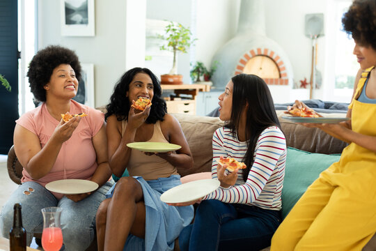 Happy young biracial female friends enjoying pizza slices while relaxing on sofa at home