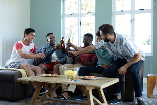 Cheerful multiracial male friends toasting beer bottles while relaxing on sofa at home, copy space