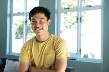 Portrait of smiling chinese young man with arms crossed standing against windows at home