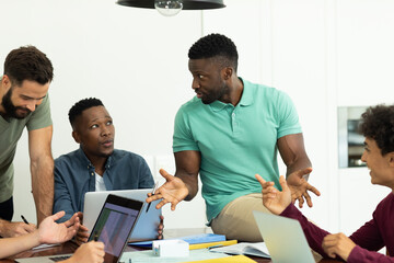 Businessman gesturing and explaining strategies to multiracial male coworkers while sitting on desk