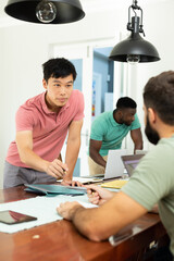 Young multiracial male entrepreneur with folders, reports on desk discussing with coworker in office