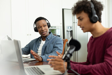 Multiracial male coworkers with headphones and laptop discussing while recording podcast in office