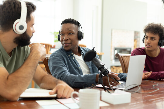 Multiracial male coworkers wearing headphones and discussing while recording podcast in office