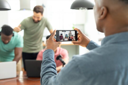 Midsection of man filming multiracial male coworkers recording podcast over mobile phone in office
