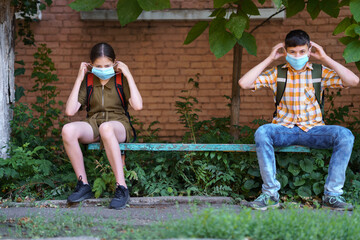 schoolchildren a boy and a teenage girl are sitting on a bench outside the school building, they use a protective breathing mask against covid or other respiratory infection