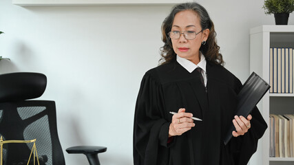 Mature female judge or lawyer or in robe gown uniform holing document and at her personal office