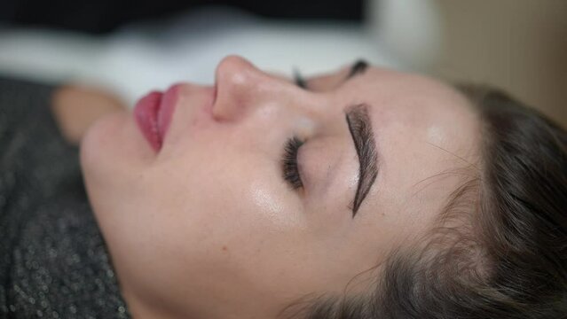 Side view close-up face of young Caucasian woman in beauty salon with hands of professional confident beautifcian contouring eyebrow. Beautiful slim client doing permanent makeup indoors