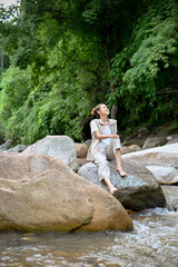 Relaxed Asian female sitting near the river on the river stone, getting some fresh air. camping