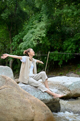 Beautiful Asian female sitting near the river on the river stone. Camping or trekking concept