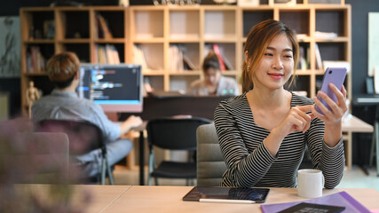 Smiling asian woman employee sitting in crate office and using mobile phone