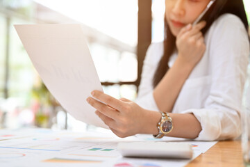 Busy adult Asian businesswoman looking some details on the paper while having a phone call