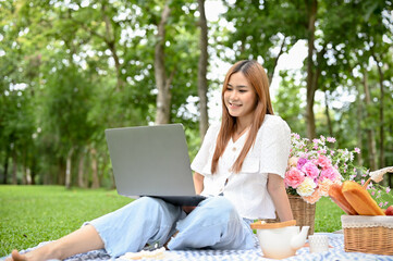 Asian female picnicking alone in the park, sitting on her picnic cloth and using laptop computer.