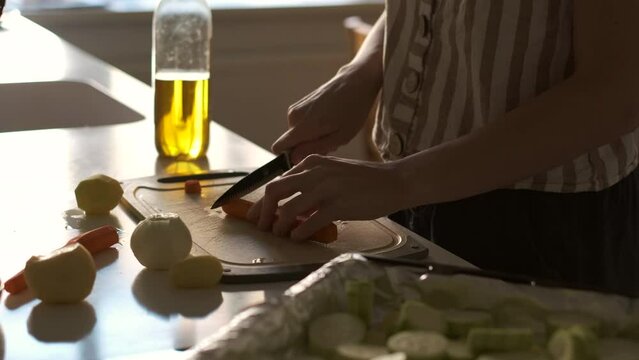 Woman slicing carrot in the kitchen preparing it to be roasted in the oven on the sheet pan. Potato, onion, olive oil and carrot are also prepared to be used in the dish