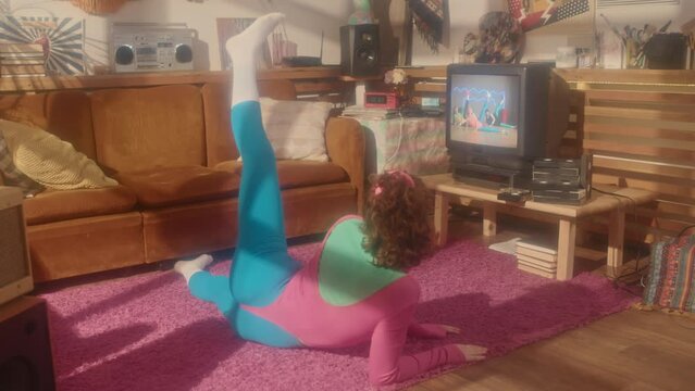 Wide rear of red-haired girl in eighties aerobic outfit exercising in living room in afternoon, watching tape on retro TV set