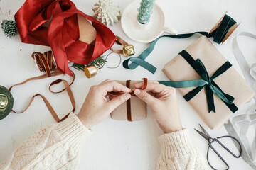 Hands wrapping stylish christmas gift flat lay. Person preparing simple christmas gift box, ribbons, craft paper and ornaments composition on white wood. Merry Christmas! Aesthetic winter holidays - obrazy, fototapety, plakaty