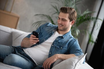 Fototapeta premium Attractive smiling young man wearing casual clothes sitting on a couch at the living room, using mobile phone