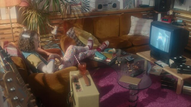 Long side view of ginger Caucasian girl sitting in armchair in room with interior design in eighties style, watching movie on retro TV and eating popcorn