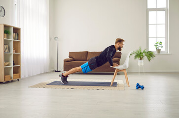 Gym at home. Athletic man is doing push up exercise with chair at home in his spacious living room with modern interior. Side view of motivated millennial guy training his muscles by doing sports.