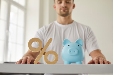 Closeup shot of percentage symbol and piggy bank on table. Close up of wooden percent sign and blue piggy bank on office desk with young man sitting in background. Money and bank interest rate concept