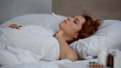redhead and sick woman measuring temperature with electronic thermometer and lying on bed.