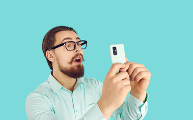 Funny astonished male in glasses reading shocking news on mobile phone on light blue background....