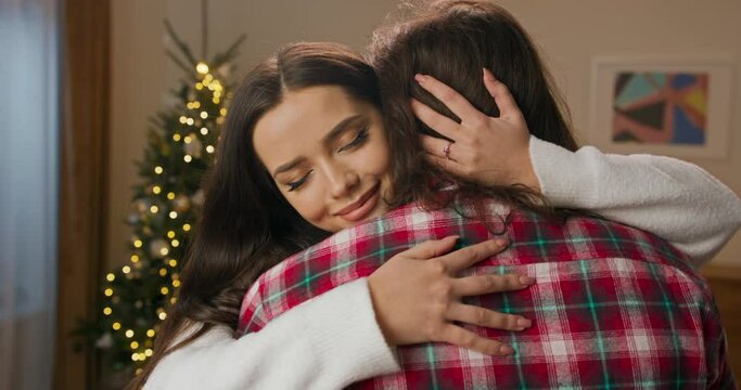 Attractive brunette girl hugs husband tightly. Young people are happy and in love. The girl is dressed in white sweater, and the guy in red checkered shirt. Behind them shimmers Christmas tree.